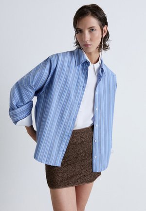 Femme portant une chemise oversize rayée bleue par-dessus un top blanc et une mini-jupe marron texturée sur un fond uni.