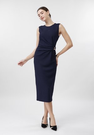 Kvinde i midikjole uden ærmer i navy med sidespændedetalje, iført sorte hæle, stående med lukkede øjne mod en ensfarvet baggrund.
