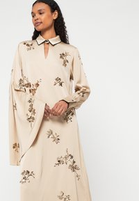 Beige színű ruha hosszú ujjal, galléros nyakkivágással és virágos flitterekkel díszítve. Sima, lágy anyag, kulcslyuk részlettel.