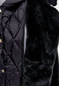 Zwarte gewatteerde vest met nepbontafwerking aan één kant. Heeft een gladde, glanzende textuur op de gewatteerde stof en een zachte, pluche feel op het bont.