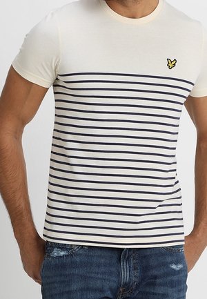Mann trägt ein weißes T-Shirt mit Rundhalsausschnitt und navyblauen horizontalen Streifen sowie einem kleinen gelben Vogel-Logo auf der Brust, kombiniert mit blauen Jeans.