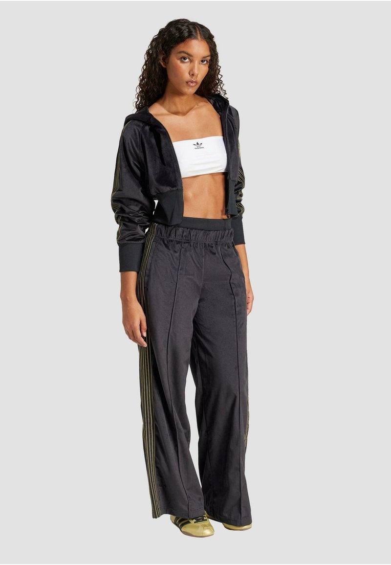 Giacca nera in velluto con zip e dettagli a strisce dorate, abbinata a un top bianco corto e pantaloni neri larghi. Scarpe metallizzate dorate completano il look.