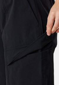 Mustat housut, joissa on sileä pinta, sivutasku napin sulkemisella. Suunnittelussa on ommeltuja saumoja ja räätälöity istuvuus.