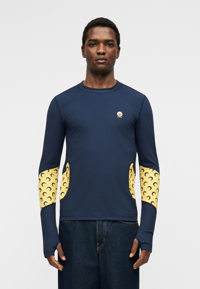 Άνδρας που φοράει μπλούζα ναυτικού μπλε με μακριά μανίκια και κίτρινες μπάλες σε σχέδιο ημισελήνου στα μανίκια και τις πλευρές, συνδυασμένη με σκούρα μπλε τζιν.