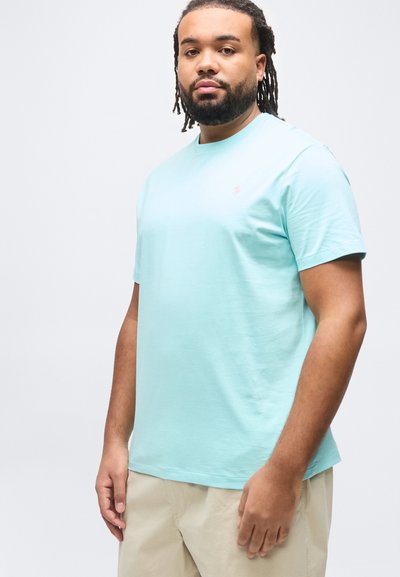 Άνδρας με γενειάδα και ντραντλοκς, ντυμένος με ανοιχτό μπλε μπλουζάκι και μπεζ παντελόνι, στέκεται μπροστά σε απλό λευκό φόντο.