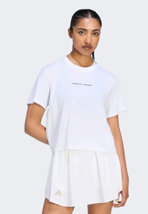 Kobieta z ciemnymi, związanymi do tyłu włosami, ubrana w biały tenisowy t-shirt Adidas i białą tenisową spódniczkę na jednolitym jasnym tle.