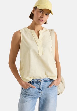 Femme portant un chemisier jaune sans manches, un jean bleu clair, une casquette jaune et un sac à dos beige, les mains dans les poches.