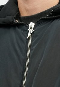 Fekete cipzáras dzseki magas gallérral, ezüst cipzárral és egyedi húzófül kialakítással. Simaszt, könnyű anyag, látható minták nélkül.