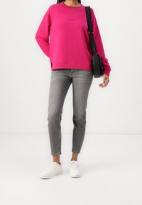 Fuksjowy sweatshirt z raglanowymi rękawami,żebrowym dekoltem i bocznymi rozcięciami. Noszony ze szarymi obcisłymi dżinsami i białymi trampkami, trzymając czarną torbę.