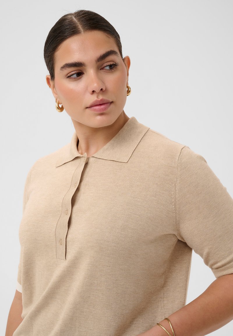 Femme aux cheveux foncés coiffés en chignon bas, portant de petites boucles d'oreilles créoles dorées, un polo beige à manches courtes et un bracelet en or au poignet droit.