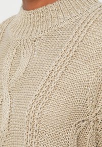 Beige színű, kábelmintás pulóver magas nyakkal. A textúrált fonal fényezése és bonyolult fonott mintázata kiemeli a pulóver eleganciáját.