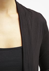 Cárdigan negro ligero con frente abierto, mangas largas y una textura suave, superpuesto sobre una camiseta de tirantes negra ajustada.