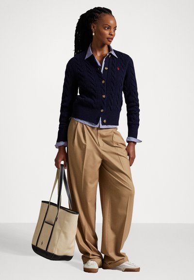 Woman with long braided hair wearing a navy cable-knit cardigan over a striped shirt, beige wide-leg pants, white sneakers, and holding a beige tote bag.