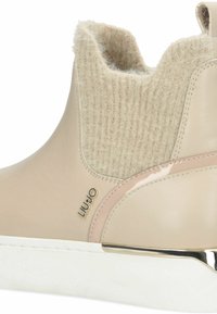 Botine bej cu parte superioară din lână texturată, accente din piele netedă, talpă albă și detaliu cu logo auriu. Design modern cu o formă rotunjită.