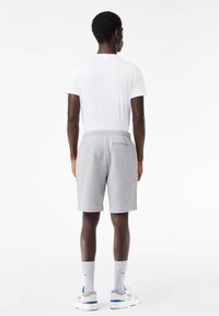 Homme debout, de dos, portant un T-shirt blanc, un short gris, des chaussettes blanches et des baskets blanches avec des accents bleus, sur un fond blanc.