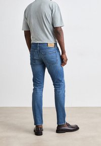 Modré džínové kalhoty s volným střihem, s kapsami na zadní straně a béžovou koženou nášivkou v pase, spárované s šedým tričkem s krátkým rukávem.