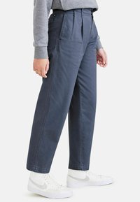 Mörkblå vida byxor med veck, gjorda av bomullsmaterial. Har ett blixtlåsstängning och en bakficka, matchat med vita sneakers.