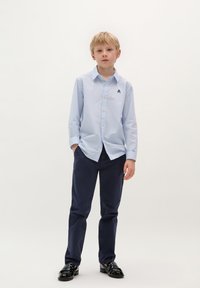 Camicia azzurra chiara a maniche lunghe con bottoni e un piccolo logo, abbinata a pantaloni blu scuro e scarpe nere lucide. Sfondo semplice.