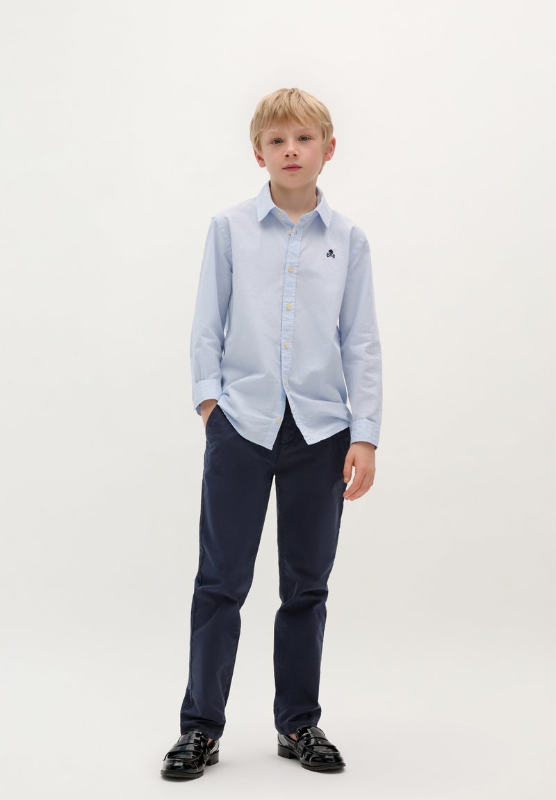 Camicia azzurra chiara a maniche lunghe con bottoni e un piccolo logo, abbinata a pantaloni blu scuro e scarpe nere lucide. Sfondo semplice.