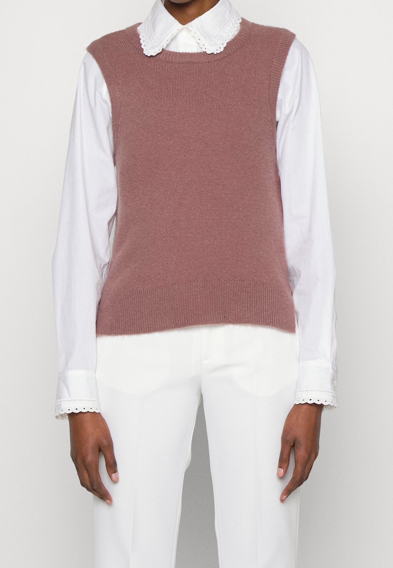 Personne portant un gilet en tricot sans manches rose poussiéreux sur un chemisier blanc à manches longues avec un col festonné et un pantalon blanc.
