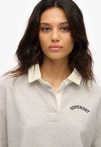 Polo gris con cuello blanco, con bordado en navy de "SUPERDRY" en el pecho, fabricado en suave material de algodón.