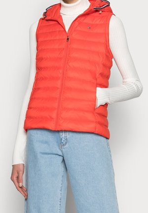 Femme portant un gilet matelassé sans manches rouge vif sur un pull à col roulé blanc côtelé et un jean bleu clair, main dans la poche du gilet.