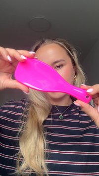 Felroze haarborstel met een plat, breed ontwerp en een textuurrijke grip; het handvat heeft een gat om op te hangen.