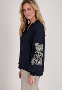 Marineblauwe blouse met een kraag en een knoopsluiting aan de voorkant. Voorzien van ingewikkeld lichtkleurig borduurwerk op de mouwen. Zachte stoftextuur.