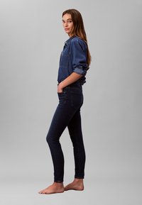 Cămașă din denim albastru închis cu mâneci rulate și buzunar, asortată cu blugi skinny albastru închis. Modelul stă desculț pe un fundal gri.