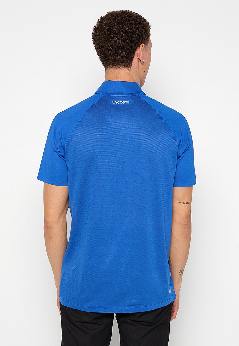 Polo bleu en tissu lisse, avec un col classique, des manches courtes et une texture subtile. Logo Lacoste au dos du cou.