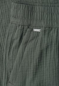 Gros plan sur un pantalon vert texturé avec une taille élastique, une poche latérale et une petite étiquette de la marque "Street One" attachée près de la poche.