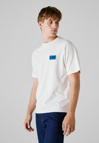 Witte katoenen T-shirt met een ronde halslijn en korte mouwen. Voorzien van een blauw "Red Bull Racing" logo op de linker borst.