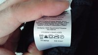 Hand houdt een wasinstructielabel vast met was symbolen en tekst in het Tsjechisch, wat aangeeft dat het 100% gerecycled polyester is, gemaakt in Bangladesh.