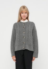 Grijs gebreide cardigan met een gestructureerd kabelontwerp, voorzien van een ronde halslijn, knoopsluiting en geribbelde manchetten. Gedragen over een overhemd met kraag.