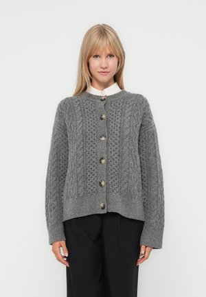 Grå strikket cardigan med et teksturert kabelmønster, med rund hals, knappelukking og ribbestrikkede ermer. Båret over en skjorte med krage.