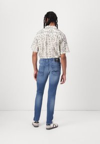 Personne aux cheveux tressés portant une chemise à manches courtes à motifs, un jean bleu et des baskets blanches, debout de dos sur un fond uni.
