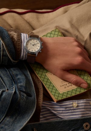 Zapestje z ročno uro Timex z usnjenim trakom, ki počiva na zeleni vintage zbirki poezije, vidna so slojena oblačila, vključno z jeans jakno in črtasto majico.