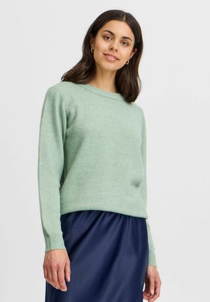 Maglione a maglia testurizzata verde chiaro con scollo rotondo e maniche lunghe, abbinato a una gonna fluida blu scuro.