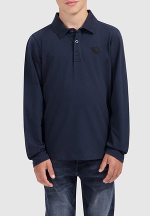 Garçon portant un polo à manches longues bleu marine avec des boutons et un jean bleu foncé, debout devant un arrière-plan uni.