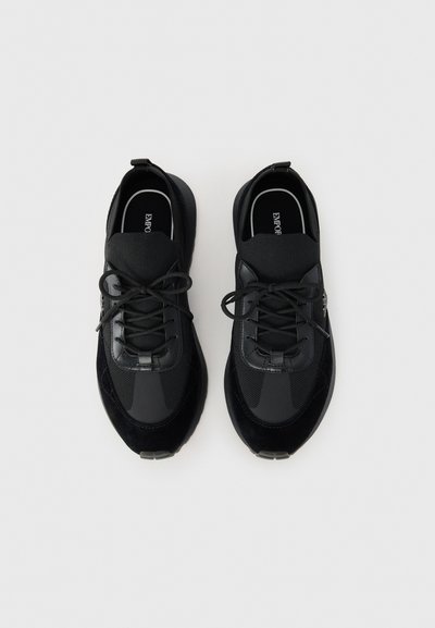Black sneakers featuring a mix of smooth leather and textured fabric. Rounded toe, lace-up design, with a pull tab at the heel.