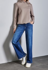 Felpa cappuccio beige e jeans a gamba larga blu, con una texture liscia e una cucitura in denim classica, abbinati a sneakers bianche.