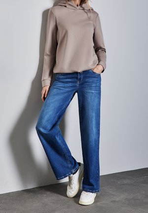 Sudadera con capucha beige y jeans de pierna ancha azules, con una textura suave y costuras clásicas de denim, combinados con zapatillas blancas.