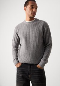 Gray ribbed knit sweater with long sleeves, crew neck, and textured fabric. Worn with dark jeans, hands positioned in pockets.