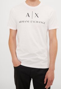 Witte katoenen t-shirt met vetgedrukte zwarte tekst "A|X ARMANI EXCHANGE", ronde halslijn en korte mouwen. Gecombineerd met donkere broek.