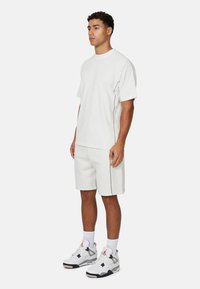 T-shirt blanc à manches courtes avec des détails de couture contrastants, associé à un short assorti. Le modèle porte des baskets blanches et grises.