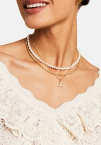Three layered necklaces: a gold chain with a Gemini pendant, a twisted gold chain, and a strand of round white pearls.