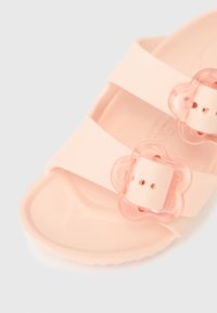 Světle růžová sandála se dvěma širokými pásky, zdobená květinově tvarovanými průhlednými přezkami. Hladký povrch a tvarovaná stélka pro pohodlí.