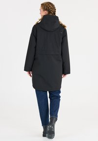 Svart vattentät parka med huva, ryggdetalj och långa ärmar. Bärs över blå jeans och svarta stövlar mot en vit bakgrund.