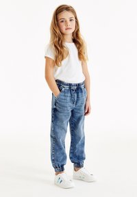 Lättvikts vit t-shirt, avslappnade blå jeansbyxor med elastisk midja och muddar, samt vita sneakers med flerfärgade detaljer.