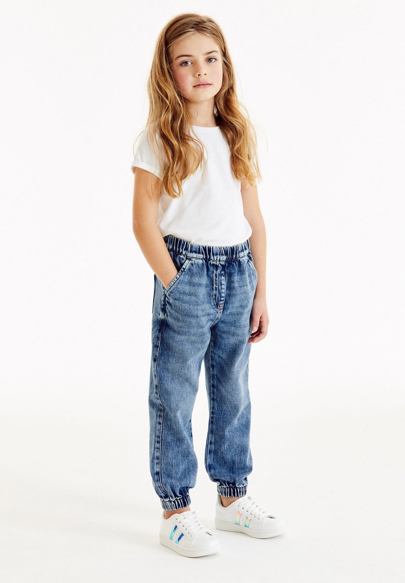 Lättvikts vit t-shirt, avslappnade blå jeansbyxor med elastisk midja och muddar, samt vita sneakers med flerfärgade detaljer.